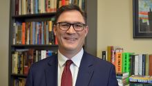 A man in a blue suit coat, white shirt, red tie wearing glasses and smiling in front of bookshelves.