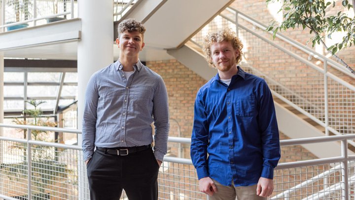 Two students stand in front of an open stairway in an atrium with lots of natural light.