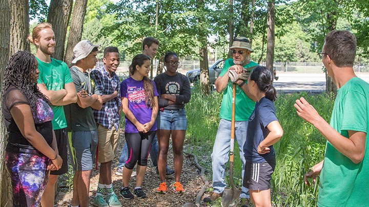 Diverse group of laughing students and a man with a shovel stand in a forest.