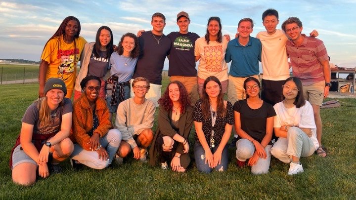 A group of 16 students in two rows stand close to one another, smile at the camera.