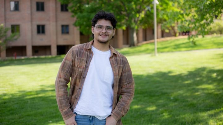 A student from Honduras in jeans and shirt wearing classes standing outside on college campus.