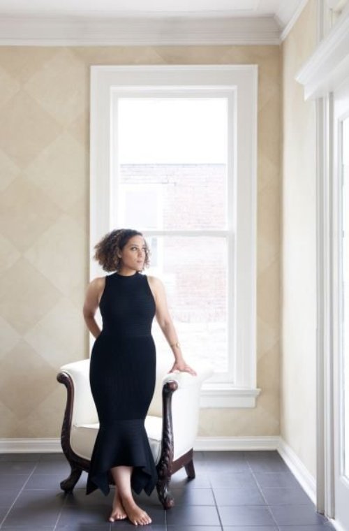 Caroline, a Black woman with chin length, curly black hair, stands by a chair wearing a long black dress, in front of a window.