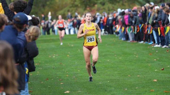 A woman in a Calvin cross country uniform runs toward the finish line past cheering fans.