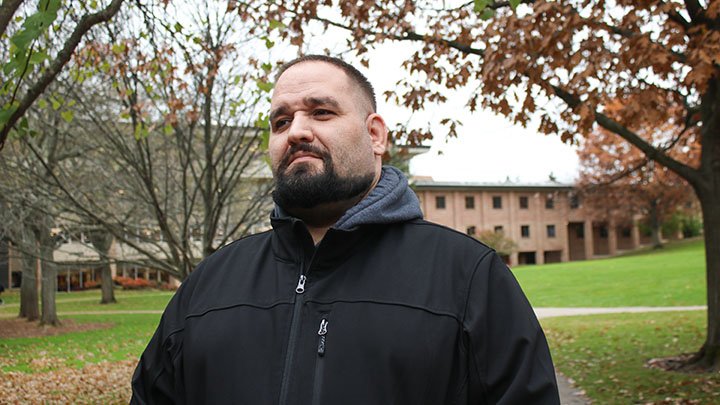 A headshot of a student in a coat looking up to the sky with campus greenery/buildings behind him.