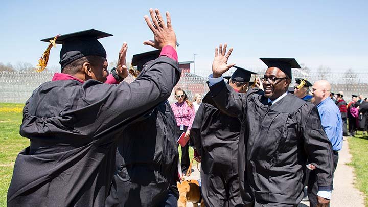 Men in caps and gowns give each other high fives outside with barbed-wire fencing in the background.