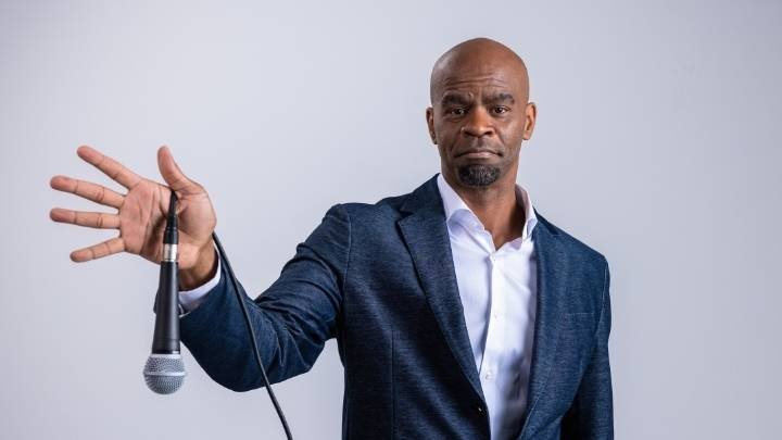 A bald African American man in a suit coat holds a mic as if he is going to drop it.