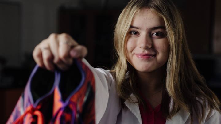 A girl in a lab coat smiles while holding a pair of track shoes in front of her.