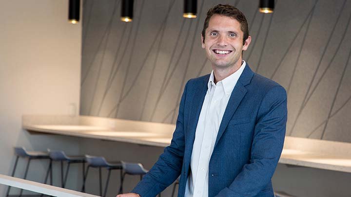 A man in a blue suit coat and white colloared shirt smiles at the camera in an indoor setting.