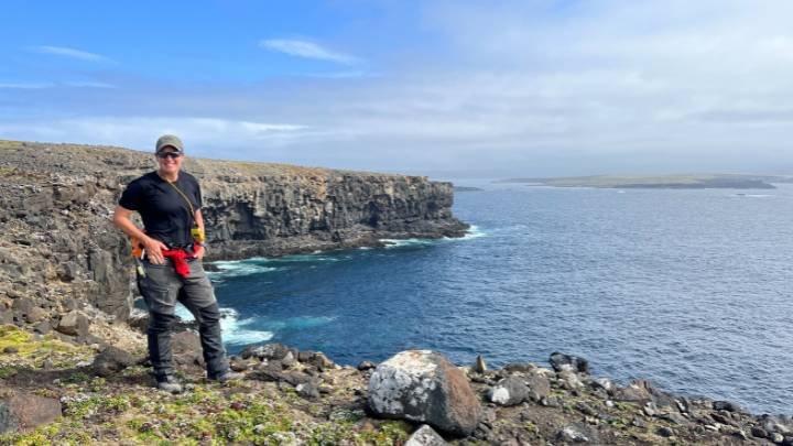 A woman wearing a T-shirt, hat, and sunglasses stands on a cliff with an ocean behind her.