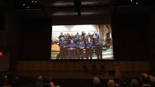 A crowd of people sit in an auditorium viewing a projection screen with inmates speaking.