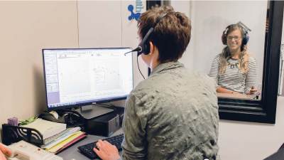 A person wearing headphones studies a computer screen with data while another looks on behind glass.