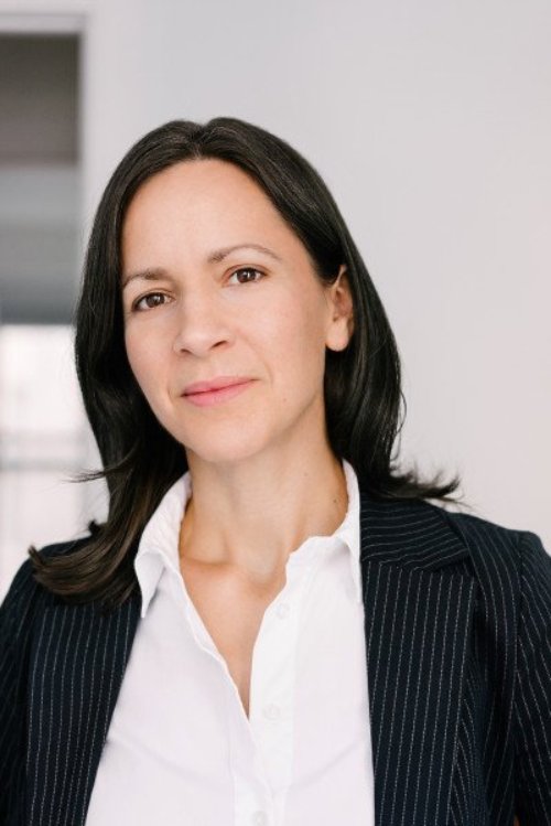 Lydia, a white woman with shoulder-length black hair, stands wearing a white-collar button up shirt and black suit jacket in front of a white wall.