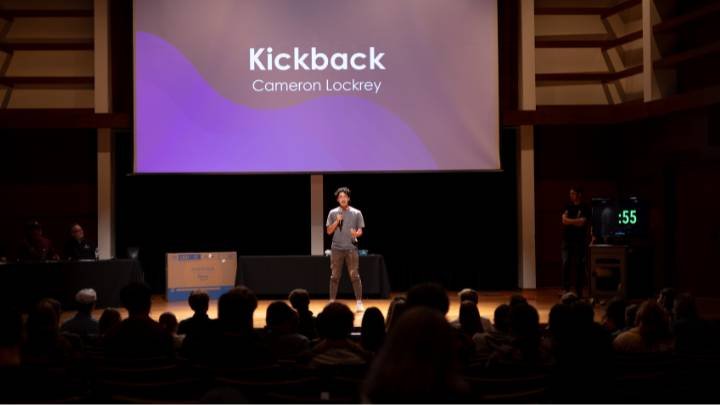 A student stands on stage under spotlight with audience looking on.