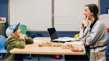 A kid in a hat holds mouth open with hands across the table from female grad student doing the same.