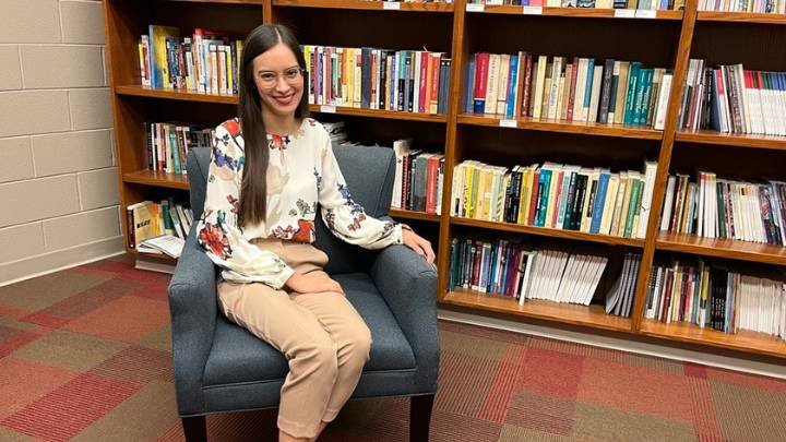 A woman sits smiles as she sits in a chair in front of a bookcase full of books.