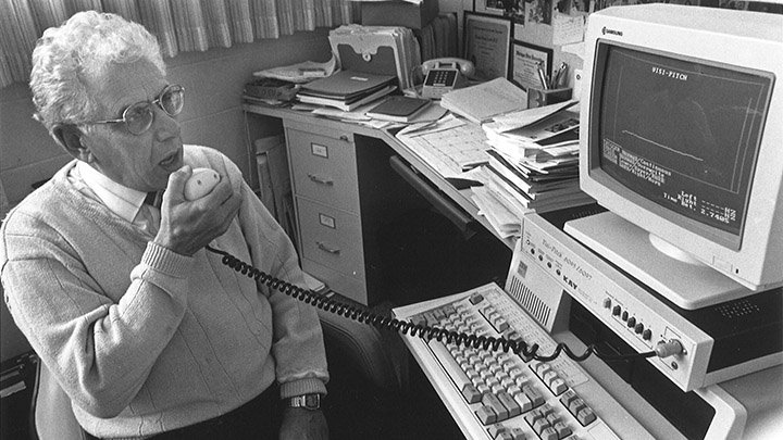 In a black and white photo, a man seated is speaking into an audiology device attached to a computer