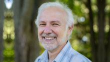 A man with white hair and a mustache and beard smiles with trees blurred behind him.