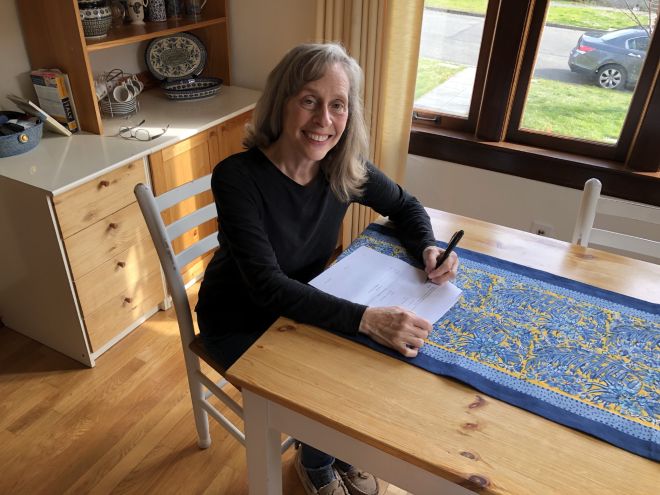 A woman seated at a kitchen table smiling as she signs a book contract.