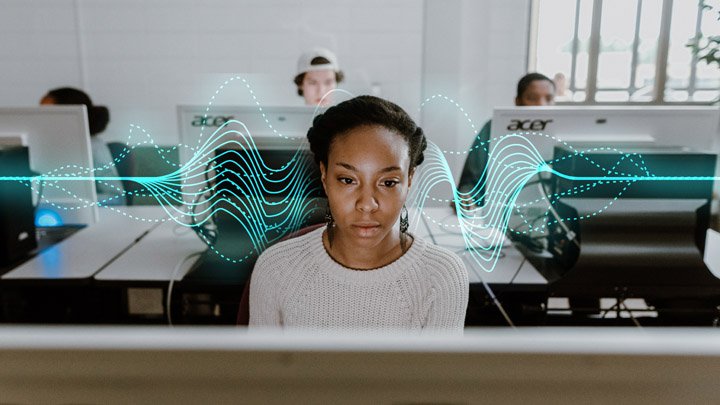 A young woman is focused on a computer in a lab with a few students seated behind her at computers.