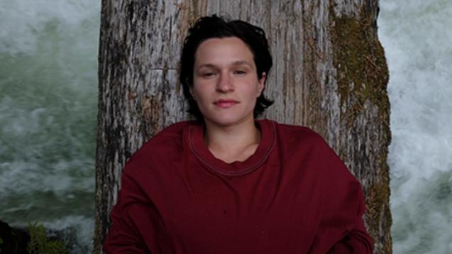 Adrianne Lenker (singer songwriter) lays on a log looking towards the camera with a half-smile.