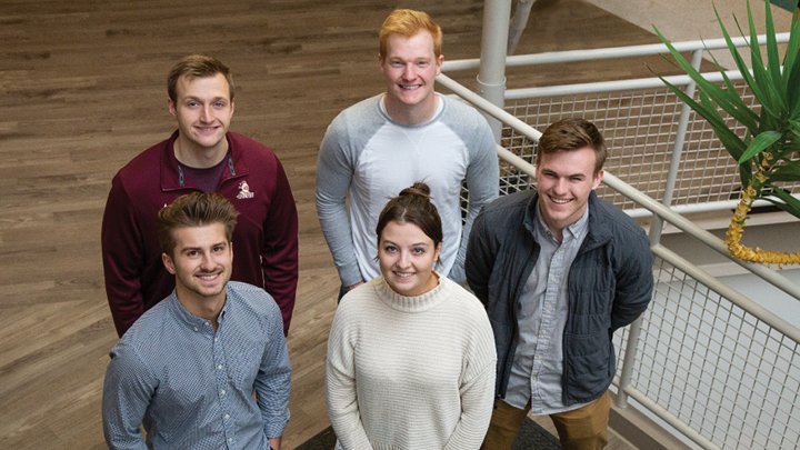 Five college students standing in an atrium looking up at the camera.