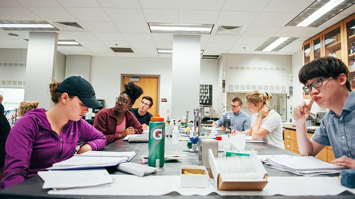 Six students listening and taking notes in a lab.