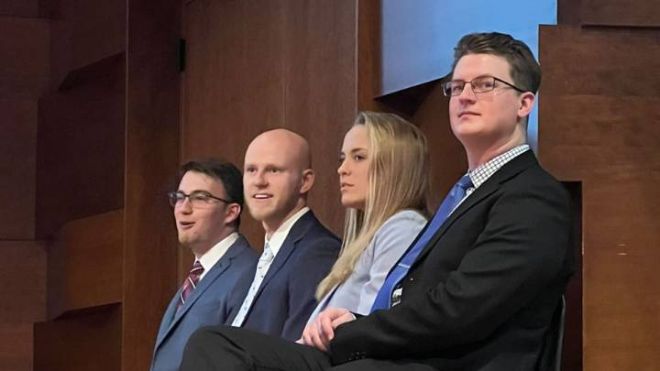 Four students (three men, one woman) in suit jackets sit as they await their presentations.