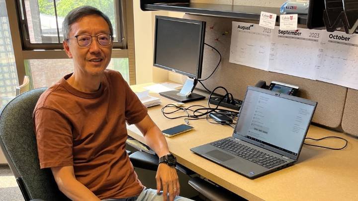 A man in a brown shirt and glasses sits at his desk smiling.