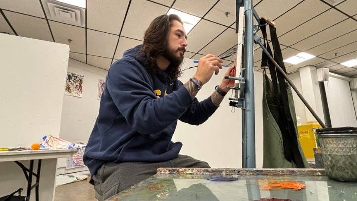 A male college student with long brown hair and a goatee paints on a canvas.