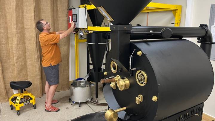 A man in an orange shirt checks an electronic device next to a coffee roaster.