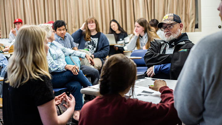 A professor, traditional college students, and adult learners sit in a circle engaged in discussion.