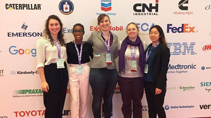 Five female students stand in front of a background filled with logos of major corporations.