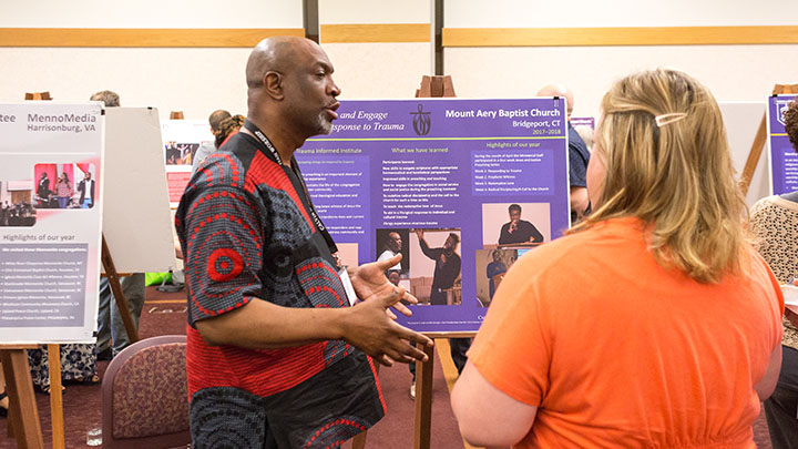 A man gestures and speaks to a woman in front of a row of posters.